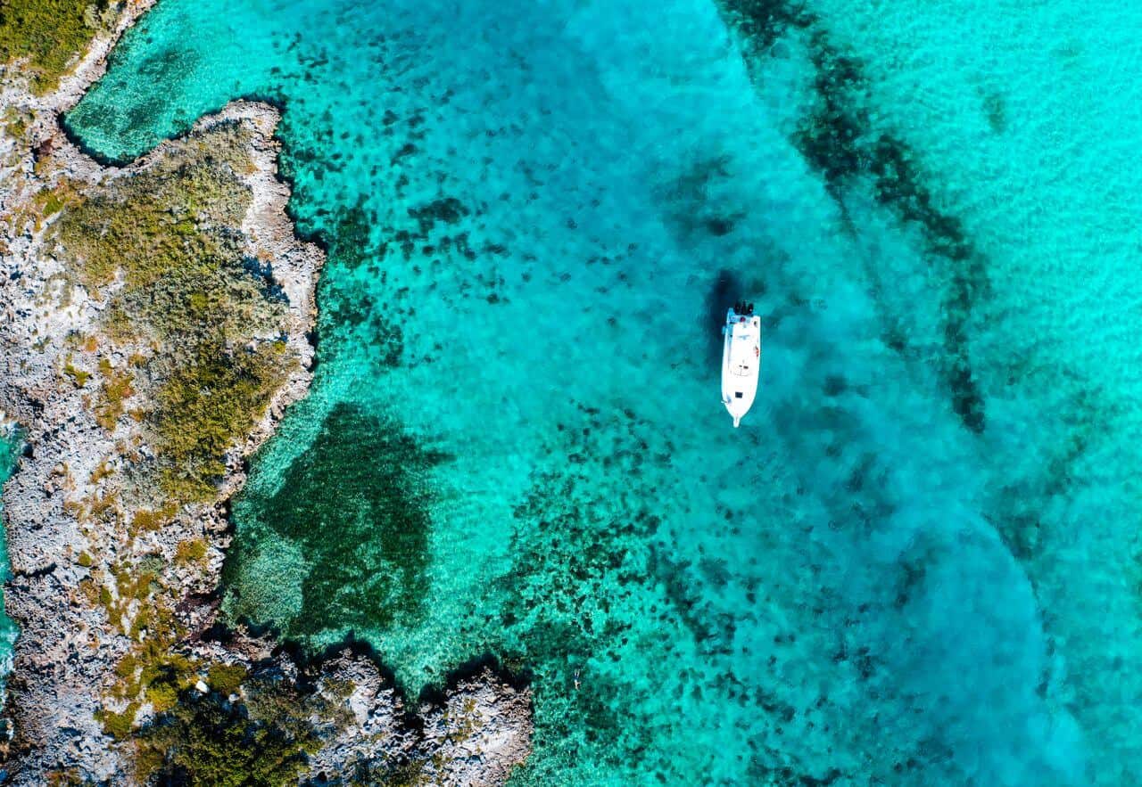 Aerial view of a white motorboat anchored over turquoise reef water beside a rocky Bahamian shoreline