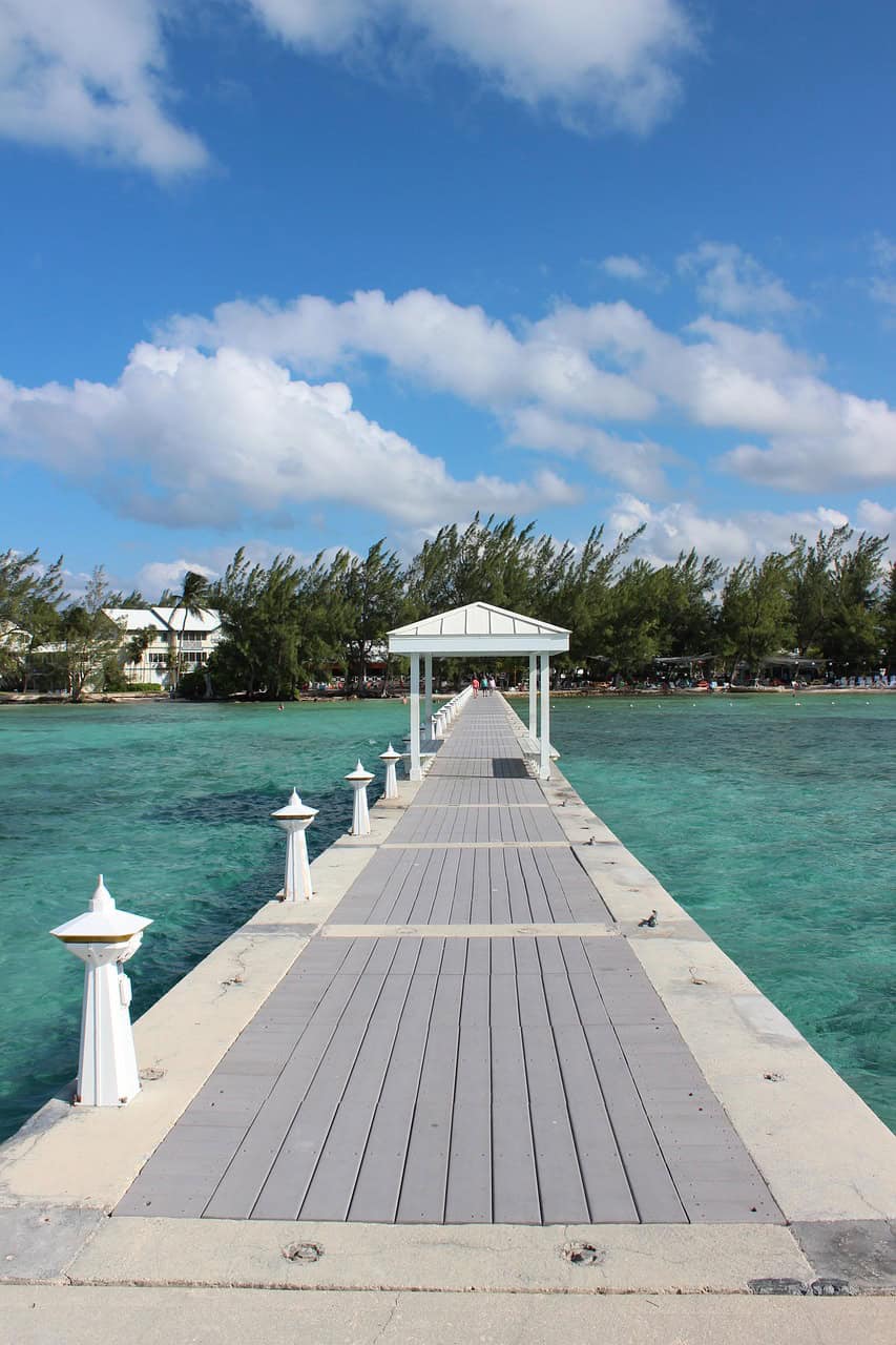 White pier with a covered gazebo extending over turquoise water towards a tree-lined shore in Grand Cayman
